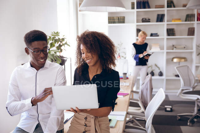 A smiling mixed race businesswoman and an African American businessman working in a modern office, using a laptop computer and talking, with their business colleagues working in the background — Stock Photo