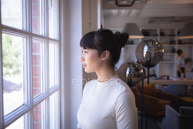 Азиатская деловая женщина, работающая в современном офисе, глядя в окно и думая, со своим коллегой, работающим на заднем плане — стоковое фото