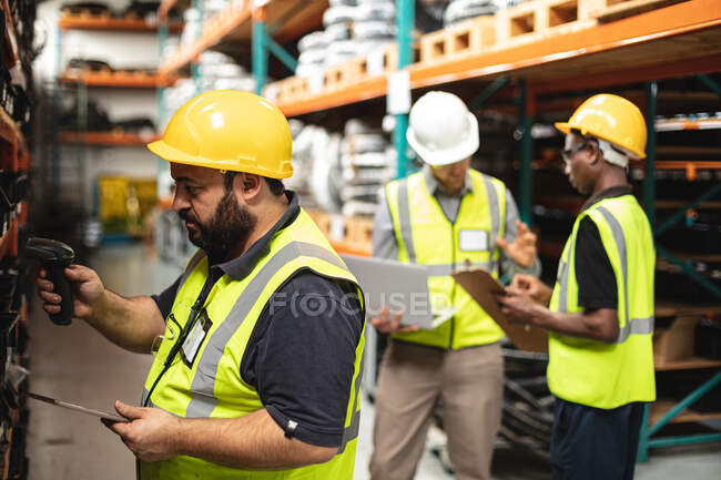 Two Caucasian and an African American male factory workers at a factory, wearing high vis vest, using laptop and checking stock. Workers in industry at a factory making hydraulic equipment. — Stock Photo