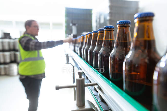 Кавказький чоловік у високій видимості, працює у мікропивоварні, перевіряє темні скляні пляшки пива на конвеєрному поясі.. — стокове фото