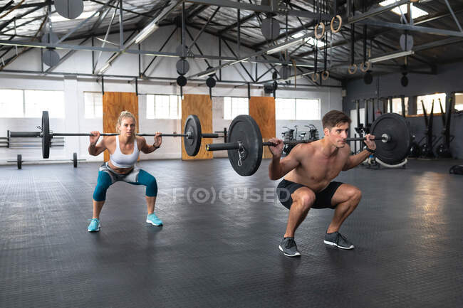 Вид сбоку атлетичный кавказский мужчина и женщина в спортивной одежде перекрещиваются в тренажерном зале, силовые тренировки с штангой, приседание на корточках с грузами на плечах — стоковое фото