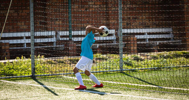 Vista lateral de un jugador de fútbol de raza mixta con su tira de equipo, en acción durante un partido de fútbol, lanzando una pelota en un campo de fútbol - foto de stock