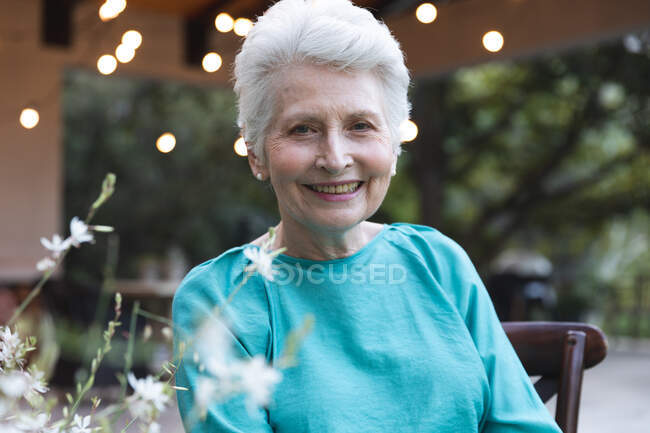 Retrato de una atractiva mujer mayor caucásica con el pelo blanco corto disfrutando de su retiro en un jardín al sol, mirando a la cámara y sonriendo, auto aislándose durante coronavirus covid19 pandemia - foto de stock