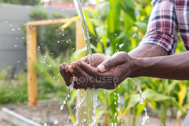 Sección media de un hombre afroamericano distanciándose en casa durante el cierre de cuarentena, lavándose las manos con un grifo de jardín. - foto de stock