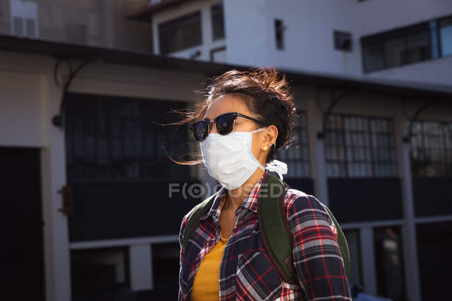 Seitenansicht einer Mischlingsfrau mit langen dunklen Haaren, die tagsüber auf den Straßen der Stadt unterwegs ist, Sonnenbrille und Gesichtsmaske gegen Luftverschmutzung und Coronavirus trägt und in einer Stadtstraße mit Gebäuden im Hintergrund spaziert. — Stockfoto