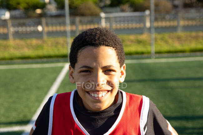 Retrato de cerca de un jugador de fútbol mixto feliz usando una tira de equipo, de pie en un campo de juego en el sol, mirando a la cámara y sonriendo - foto de stock