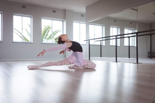 Bailarina feminina caucasiana atraente com cabelo ruivo que se estende para trás, preparando-se para uma aula de balé em um estúdio brilhante, com foco em seu exercício, sentada no chão . — Fotografia de Stock