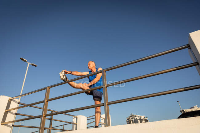 Niedriger Winkel Seitenansicht eines älteren kaukasischen Mannes, der an einem sonnigen Tag mit blauem Himmel auf einer Promenade trainiert und sich mit einem Bein auf einer Brüstung ausstreckt — Stockfoto
