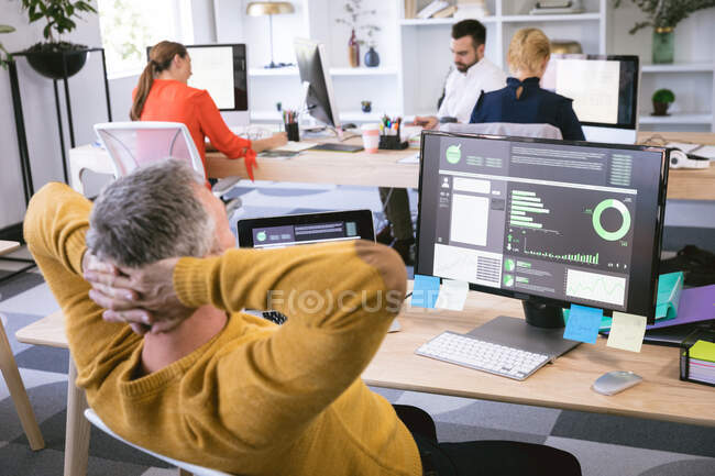 Un uomo d'affari caucasico che lavora in un ufficio moderno, seduto a una scrivania con le mani dietro la testa guardando uno schermo del computer, con i suoi colleghi di lavoro che lavorano sullo sfondo — Foto stock