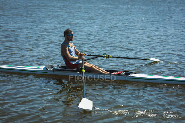Бічний вид на кавказького веслувальника, який тренується і веслує на річці, тримаючи весла і сидячи в веслувальному човні в сонячний день. — стокове фото