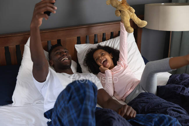 Menina afro-americana e seu pai social distanciamento em casa durante o bloqueio de quarentena, passar o tempo juntos, se divertindo e tirar uma selfie com um smartphone . — Fotografia de Stock