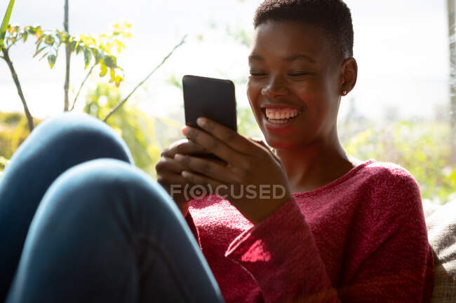 Vue de face gros plan d'une Afro-Américaine assise dans son salon devant une fenêtre par une journée ensoleillée, à l'aide d'un smartphone et souriant — Photo de stock