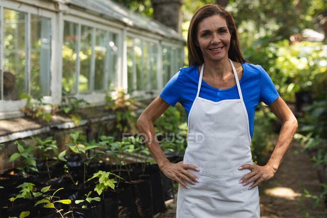 Portrait a Caucasian woman with long brown hair wearing an apron, enjoying time in a sunny garden, looking at camera and smiling — Stock Photo