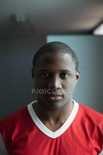 Portrait close up of a teenage African American male field hockey player, wearing a red team strip, standing in the changing room, looking straight to camera, focusing before a game — Stock Photo