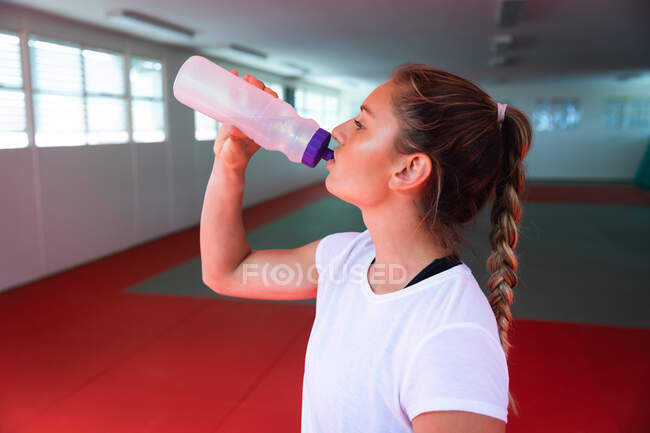 Vue latérale rapprochée d'une judoka femme caucasienne buvant de l'eau à partir d'une bouteille en plastique, debout dans la salle de gym, faisant une pause dans l'entraînement. — Photo de stock