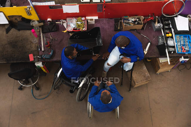 Grupo de trabajadores afroamericanos discapacitados en un taller en una fábrica que fabrica sillas de ruedas, sentados en un banco de trabajo ensamblando partes de un producto, dos sentados en sillas de ruedas, uno usando muletas - foto de stock