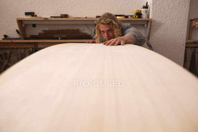 Creatore di tavole da surf caucasiche con lunghi capelli biondi, che lavora nel suo studio, fa una tavola da surf, la ispeziona e si prepara alla lucidatura... — Foto stock