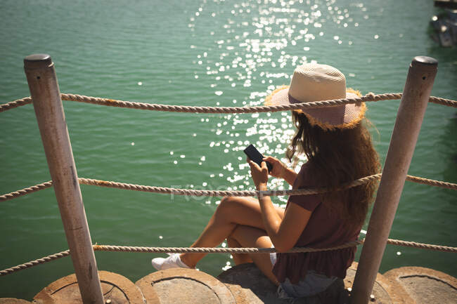 Ein kaukasisches Mädchen im Teenageralter mit Strohhut genießt an einem sonnigen Tag die Zeit auf einer Promenade, im Sitzen und mit einem Smartphone — Stockfoto