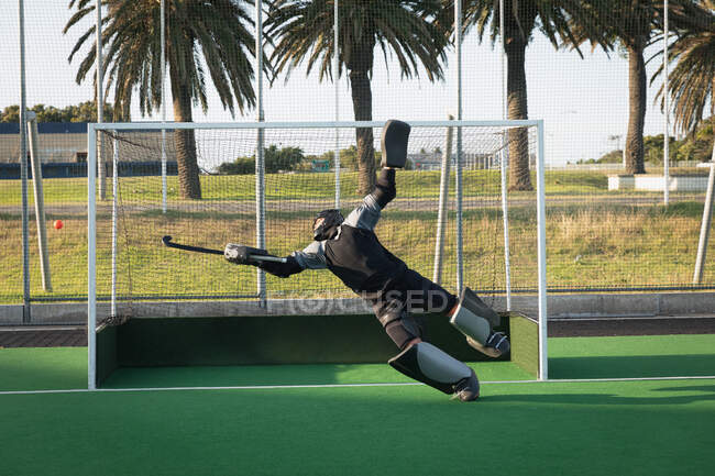 Front view of a teenage Caucasian male field hockey goalkeeper in action on the pitch, diving to stop a ball with a hockey stick, during a hockey match between two multi-ethnic teenage male teams on a sunny day — Stock Photo