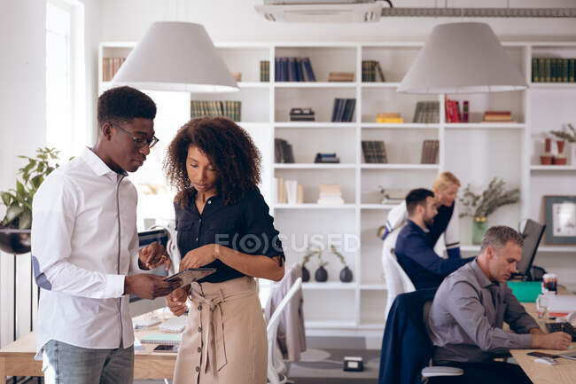 Uma mulher de negócios mista e um empresário afro-americano trabalhando em um escritório moderno, usando um tablet e conversando, com seus colegas de negócios trabalhando em segundo plano — Fotografia de Stock