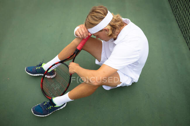 Ein kaukasischer Mann in weißen Tennisanzügen, der an einem sonnigen Tag auf einem Tennisplatz sitzt und einen Tennisschläger in der Hand hält — Stockfoto