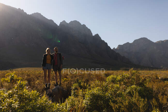 Vorderansicht eines kaukasischen Paares, das eine gute Zeit auf einer Reise in die Berge hat, auf einem Feld unter den Bergen spazieren geht, zusammen auf einem Felsen steht, an einem sonnigen Tag — Stockfoto