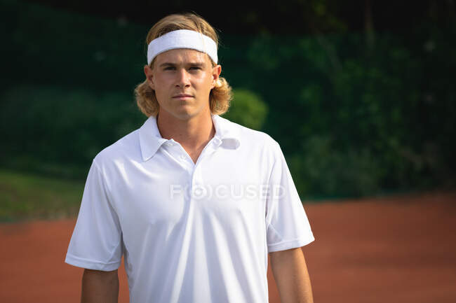 Ritratto di un uomo caucasico che indossa dei bianchi da tennis che passa del tempo su un campo a giocare a tennis in una giornata di sole, guardando la macchina fotografica — Foto stock