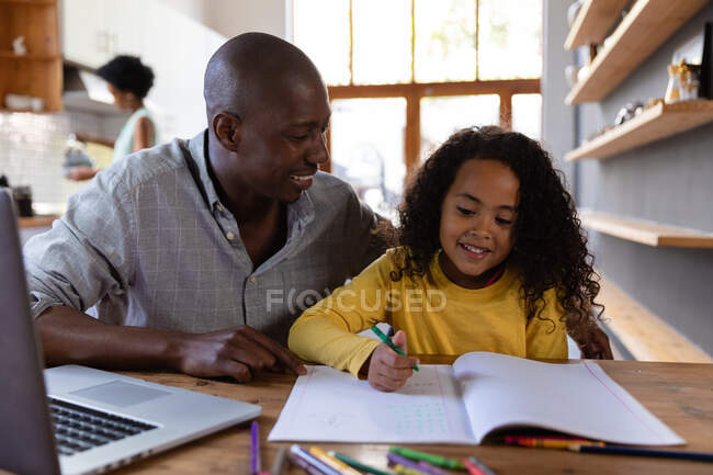 Vista frontal de cerca de un hombre afroamericano en casa, sentado en una mesa con su hija pequeña mirándola dibujar en un libro escolar y ambos sonriendo, una computadora portátil en la mesa frente a él - foto de stock