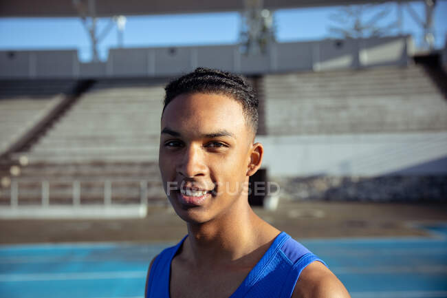 Портрет впевненого змішаної раси спортсмена, який практикує на спортивному стадіоні в синьому жилеті, дивлячись на камеру і посміхаючись — стокове фото