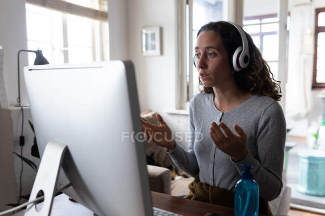 Femme caucasienne passant du temps à la maison, portant des écouteurs, assise à son bureau et travaillant à l'aide de son ordinateur. Distance sociale et isolement personnel en quarantaine. — Photo de stock