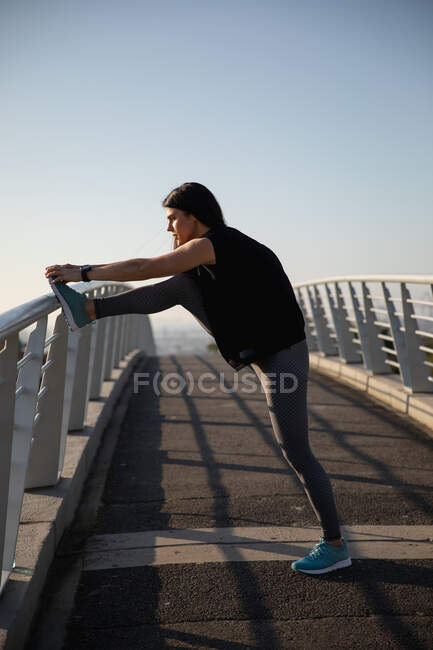 Vista lateral de una mujer caucásica en forma con el pelo largo y oscuro usando ropa deportiva haciendo ejercicio al aire libre en la ciudad en un día soleado con cielo azul, calentándose, estirando su pierna, - foto de stock