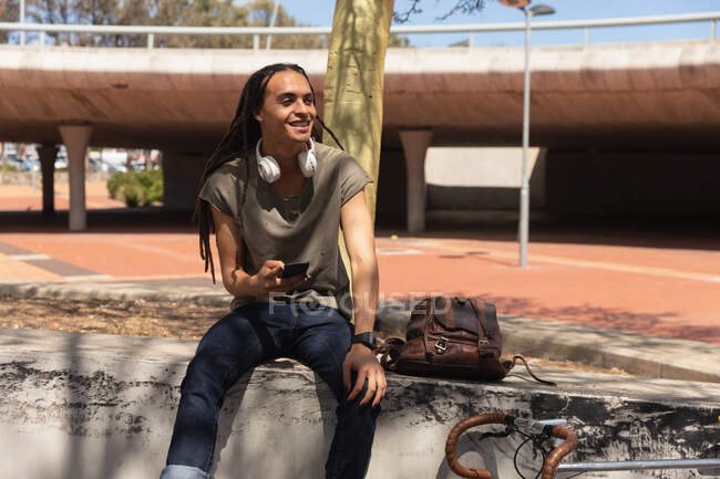 Side view of a mixed race man with long dreadlocks out and about in the city on a sunny day, sitting on a wall in the street and smiling, using a smartphone, with his bicycle leaning against the wall next to him. — Stock Photo
