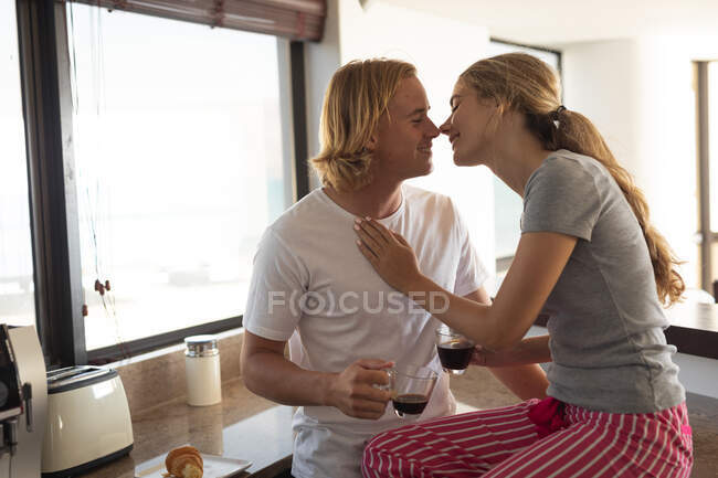 Casal caucasiano em pé em uma cozinha, bebendo café, abraçando e beijando. Distanciamento social e auto-isolamento em quarentena. — Fotografia de Stock