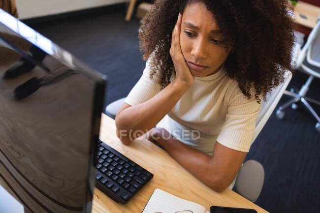 Втомлена змішана гоночна бізнес-леді з кучерявим волоссям, працює в сучасному офісі, сидить за столом, використовуючи настільний комп'ютер — стокове фото