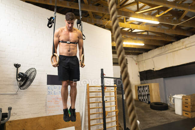 Vista frontal de um homem atlético caucasiano sem camisa usando uma cinta torácica monitor de frequência cardíaca treinamento cruzado em um ginásio, levantando-se em anéis de ginástica — Fotografia de Stock