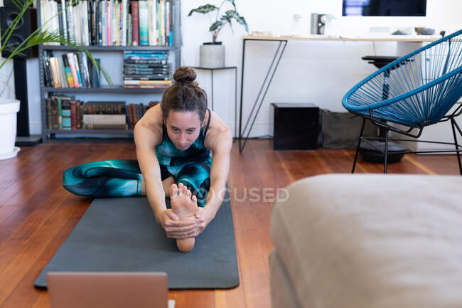 Caucasian woman spending time at home, wearing sportswear, exercising on a mat, joining online yoga course, using her laptop. Social distancing and self isolation in quarantine lockdown. — Stock Photo
