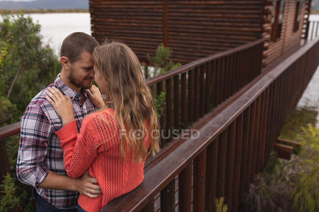 Vista lateral de um casal caucasiano se divertindo em uma viagem às montanhas, de pé em uma varanda em uma cabine, abraçando, tocando suas cabeças juntas — Fotografia de Stock