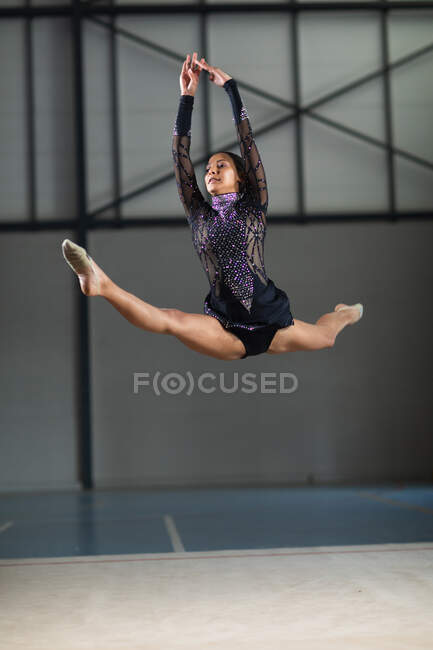 Vista frontal de la gimnasta femenina adolescente de raza mixta que actúa en el gimnasio, saltando y haciendo split, vistiendo un maillot negro y púrpura - foto de stock