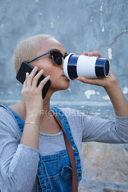 Mixed Race Alternative Frau mit kurzen blonden Haaren unterwegs in der Stadt an einem sonnigen Tag, mit Smartphone, Sonnenbrille und Kaffee zum Mitnehmen. Urbaner digitaler Nomade unterwegs. — Stockfoto