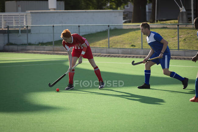 Side view of two teenage Caucasian male field hockey players from opposing teams in action on the pitch during a hockey game on a sunny day — Stock Photo
