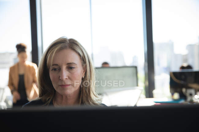 Eine kaukasische Geschäftsfrau, die in einem modernen Büro arbeitet, am Schreibtisch sitzt und einen Computer benutzt, während ihre Kollegen im Hintergrund arbeiten — Stockfoto