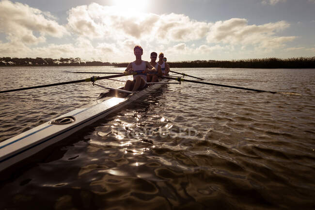 Vista frontal de ángulo bajo de un equipo de remo de cuatro hombres caucásicos entrenando y remando en el río, sentados en un bote de remo al atardecer - foto de stock