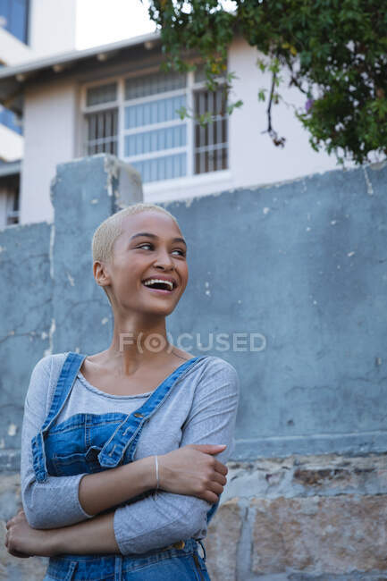 Femme alternative de race mixte aux cheveux blonds courts portant des salopettes en denim, en ville par une journée ensoleillée, regardant ailleurs et riant. Femme indépendante urbaine en déplacement. — Photo de stock