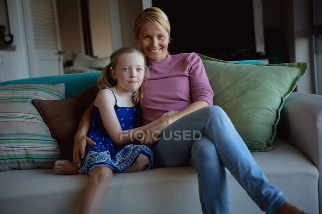 Портрет белой женщины, наслаждающейся семейным отдыхом с дочерью дома вместе, сидящей на диване в гостиной и обнимающей друг друга, улыбающейся в камеру — стоковое фото