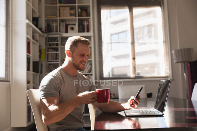 Side view mid section of a young Caucasian man spending time at home, sitting by the desk, using his laptop computer and smartphone and holding a cup of hot beverage. — Stock Photo