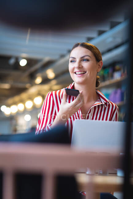 Una mujer de negocios caucásica con el pelo corto, sentada en una mesa dentro de un café, hablando en su teléfono inteligente y sonriendo, vistiendo ropa de moda - foto de stock