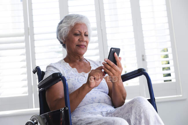 Une femme afro-américaine retraitée âgée à la maison, assise dans un fauteuil roulant portant un pyjama devant une fenêtre par une journée ensoleillée à l'aide d'un smartphone et souriante, s'isolant elle-même pendant une pandémie de coronavirus covid19 — Photo de stock