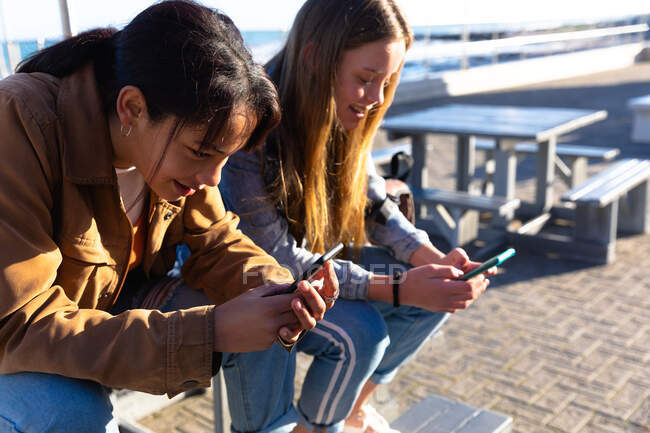 Vista lateral de un caucásico y una raza mixta niñas disfrutando del tiempo juntos en un día soleado, sentado en un banco en el paseo marítimo, sosteniendo y utilizando sus teléfonos inteligentes. - foto de stock
