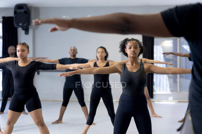 Front view of a multi-ethnic group of fit male and female modern dancers wearing black outfits practicing a dance routine during a dance class in a bright studio, spreading their arms. — Stock Photo