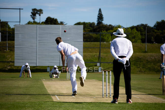 Rückansicht eines jugendlichen kaukasischen Cricketspielers in weißen Kleidern, der den Ball während eines Cricketspiels auf das Spielfeld wirft, während ein Schiedsrichter hinter ihm steht. — Stockfoto